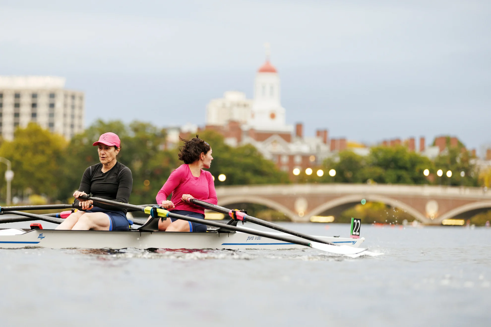 Two rowers head towards the John W. Weeks Footbridge with Dunster House visible in the background.