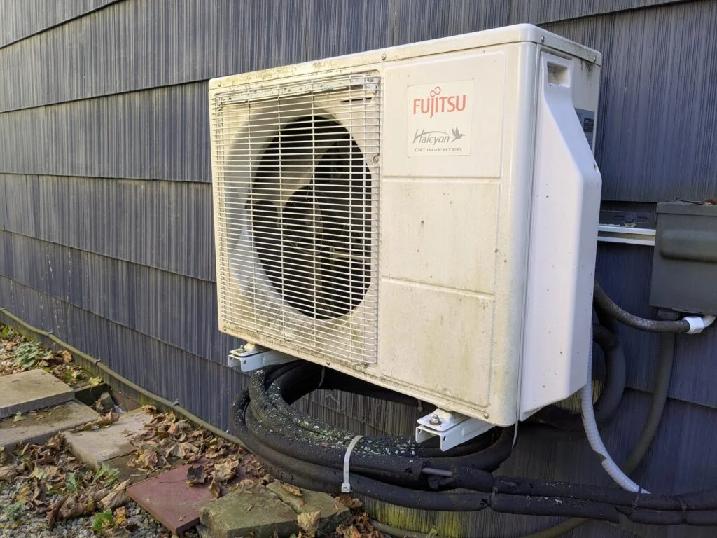 A heat pump is mounted to an exterior wall of a blue house. The pump is a gray rectangular prism about the size of a sofa that says "Fujitsu Halcyon." Roughly two-thirds of the front face of the heat pump is taken up by a fan. Black tubes and cables run from the bottom of the pump into the house.